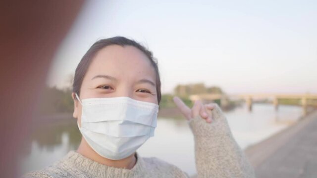 Close up of happy Asian woman standing and taking video selfie by the river. wearing mask. River and trees background, looking beautiful view