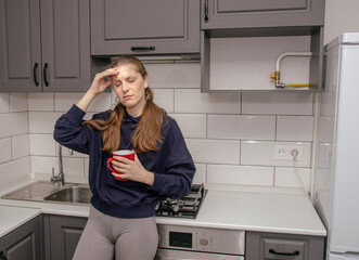 A young brunette girl stands sad and unhappy in the kitchen alone with a red mug, holds her right hand to her head because of a headache