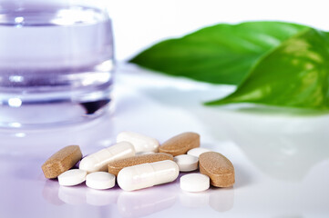 white pills and caplsules, glass of water and green leafs on white background close up