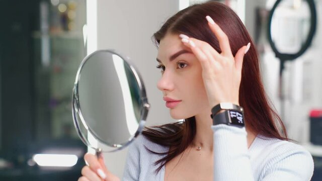 Beautiful young woman with perfect skin face looks at herself in the mirror in the beauty salon. Preparation for permanent eyebrow makeup. Cosmetology procedures.