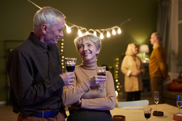 Beautiful aged spouses with red wine glasses in hands standing in decorated with electric lights living room and talking