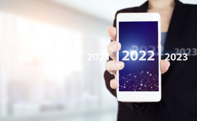 New year concept. Hand hold white smartphone with digital hologram 2022 sign on light blurred background. 2022 new year. Year two thousand and twenty two concept.
