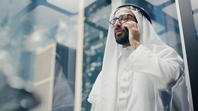 Successful Muslim Businessman in Traditional White Kandura Riding Glass Elevator to Office in Modern Business Center. Man Talking on a Phone in a Lift. Saudi, Emirati, Arab Businessman Concept.