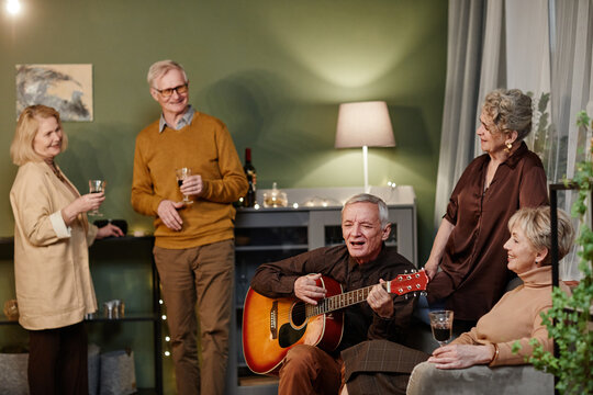 Talented aged Caucasian man playing acoustic guitar and singing during meeting with friends. Four seniors listening to their fried performance