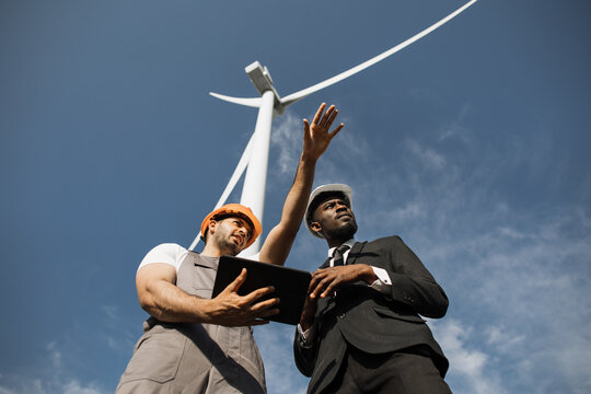 Indian engineer in uniform and helmet using tablet during meeting with african american inspector in stylish suit. Two partners discussing green energy while standing on farm with windmills.
