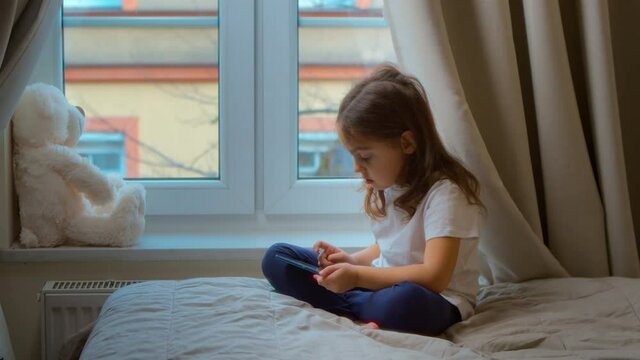 Cute little girl is sitting on a cozy bed in the bedroom by the window, using a smartphone, playing video games, watching cartoons, spending time on weekends with a modern technological concept.