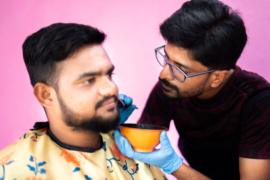 focus on barber, close up shot of concentrated hairdresser applying hair dye or black henna to young man to cover white hair beard at salon - concept of hair coloring service and small business.