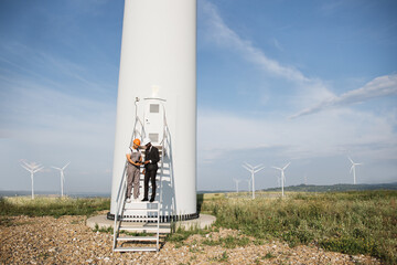 Multiracial partners standing on stairs near wind turbine and looking on screen of digital tablet. Two industrial workers turning on system operation of eco farm using modern gadgets. © sofiko14