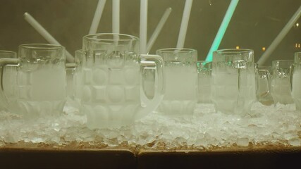 Row of many empty frozen beer glasses . 0.3 l and 0.5 l cups of beer on table . Neon green flashing lights background . Ice cubes on table . Slow motion . Shot on ARRI ALEXA Camera with Laowa lens