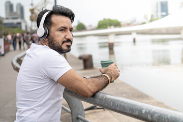 Fototapeta na wymiar Latin man with a beard, headphones and a mug of coffee looking at the river.
