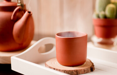 clay ceramic mug, hot with steam on white wooden tray and tea or coffee pot with rustic wooden background