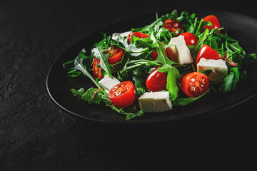 Fresh salad with tomatoes, arugula and cheese cubes on black plate on Dark grey black slate background