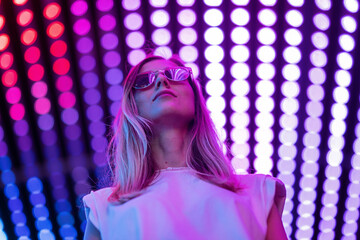 Teen hipster girl in stylish glasses standing on purple tunnel with neon light wall background,...