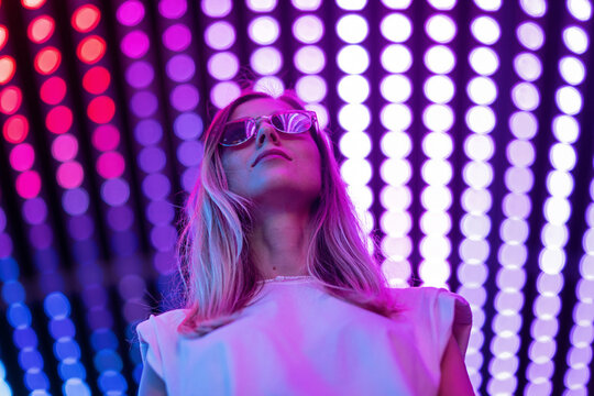 Teen Hipster Girl In Stylish Glasses Standing On Purple Tunnel With Neon Light Wall Background, Female Teenager Fashion Model Pretty Young Woman Looking At Night Club City Light Glow