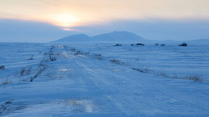 A road among the snow-covered tundra in the Arctic. View of the road, old abandoned houses in the tundra and snow-capped mountains. Winter Arctic landscape. Travel to the Far North of Russia. Chukotka