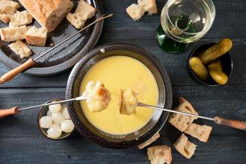 Swiss cheese fondue as new year party meal with bread on long forks, pickles and wine on a dark wooden table, high angle view from above, selected focus