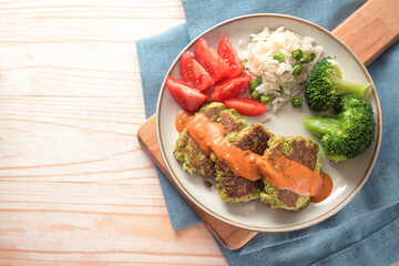 Vegetable patties with broccoli, tomato, rice and a hot sauce, vegetarian and vegan meal on a plate and a cutting board, blue napkin and light wooden table, top view from above, copy space,