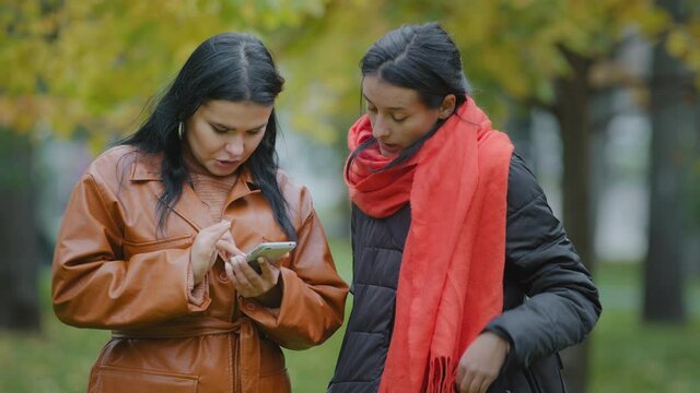 Two hispanic girls stand in autumn park using telephone women ordering food taxi in mobile application watching photos on dating site on social networks on smartphone choosing clothes in online store