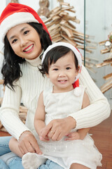Portrait of cheerful mother in Santa hat and cute little daughter sitting on her laps