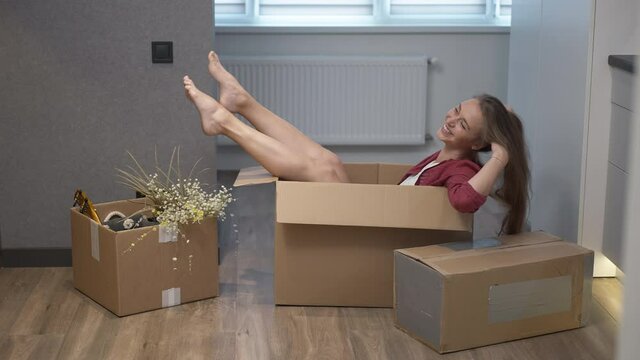 Wide shot happy female apartment owner lying in cardboard box shaking legs in slow motion smiling. Side view excited successful rich Caucasian millennial woman enjoying relocation indoors
