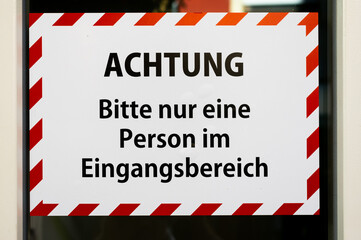Schild „Achtung Bitte nur eine Person im Eingangsbereich“ in Deutsch mit weiß rotem Rand wie Apsperrband wegen Corona an einer Eingangstür in Großaufnahme bildfüllend