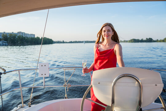 The pritty woman in a long red dress holding a glass of champagne standing on a yacht and looking for river sunset view