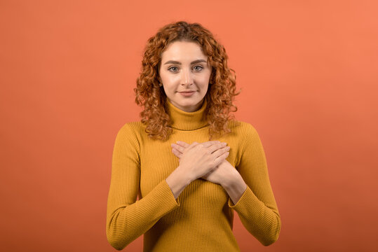 Young and attractive redhead Caucasian girl in orange jumper showing gratitude gesture while holding hands at chest isolated on orange studio background.