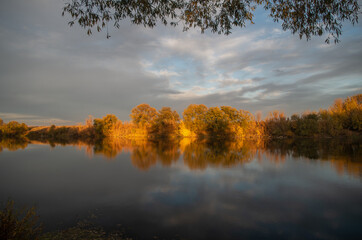 autumn trees on the shore of the lake with a reflection against the background of a stormy sky and the setting sun