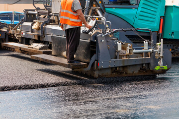 A road worker at the control panel of a tracked paver paver paving fresh asphalt on a fenced road on a summer day.