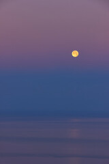 foto vertical de una luna llena sobre el mar con unos colores magentas y violetas por toda la escena, el mar esta en calma y tiene un pequeño reflejo de la luna