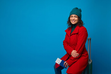 Delightful woman in red coat, green hat and pullover, with passport, air ticket, boarding pass, sits on suitcase and smiles toothy smile looking at camera. Travel. Tourism concept. Blue background