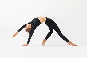 Fototapeta na wymiar A young woman practicing yoga performs the exercise of Kamatkarasana, the pose of a dancing dog, trains in black sportswear on a white background