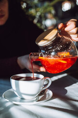 The hand of unrecognizable woman pours hot tea from glass kettle to mug sitting by table with white tablecloth.