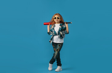 a little hooligan girl in torn jeans with a baseball bat in her hands on an isolated blue...
