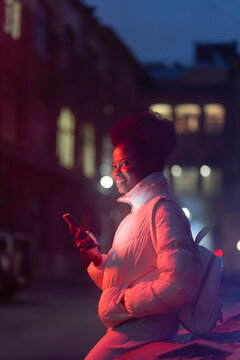 Young African Woman At Night With Smartphone Wait For Taxi From Booking Mobile App. Black Millennial Female Use Cellphone Outdoors Looking For Blind Date From Dating Application. Technology And Gadget