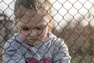 A little girl with a sad look behind a metal fence. Social problem of refugees and forced migrants