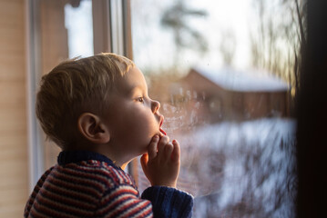 little boy stand near the window and look at the snowy street in winter, when it is very cold