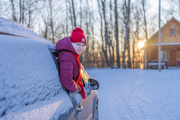Fototapeta na wymiar young woman in a red hat peeking out of a snow-covered car on the background of a winter road and the setting sun.