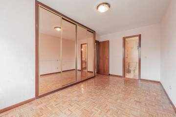 Empty bedroom with white walls with a small slatted parquet floor, a large built-in wardrobe with sliding mirrored doors and exit to the side of the bathroom