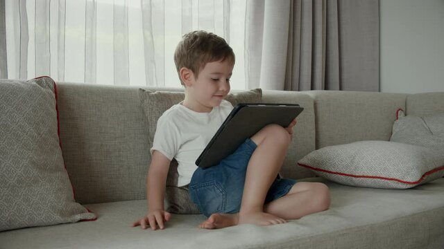 Children tech addiction concept. Child with gadget.Cute preschool kid boy using digital tablet technology device online education sitting on couch at home.
