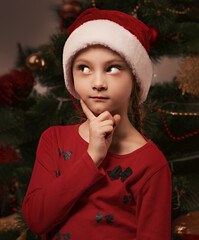 Cute kid girl with in Christmas santa claus hat thinking about new year gift and holding the finger under the face on green fur tree background. Closeup portrait