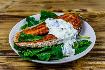 Plate with roasted salmon steak, tartar sauce and spinach leaves on a wooden table