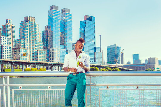 Waiting for You. Wearing a white, long sleeve shirt,  green pants, a young black guy with mohawk hair is standing in the front of buildings, hands holding a white rose, looking at you. City Love.