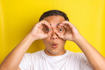 Close-up portrait of amazed Asian man with gesture pretending to make binoculars with his hands isolated on yellow background