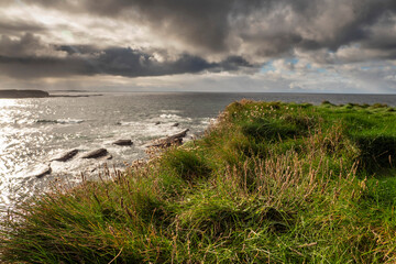Fototapeta na wymiar Green grass on a cliff and rough stone coastline. Dark dramatic cloudy sky. West of Ireland. Irish landscape. Nature scene.