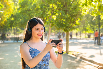 Young woman at park using a dress during summer spring season at a sunny day. Brunette with long black hair smiling using a smart phone and headphones