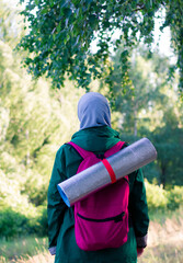 A Muslim woman in a green jacket and a blue hijab with a backpack and a hiking mat in the forest. Hiking in the mountains.