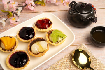 Homemade Fruits tarts, Sweet desserts topping with fresh kiwi, blueberries, peach, longan, raspberries in syrup, served with hot herbal tea on wooden table