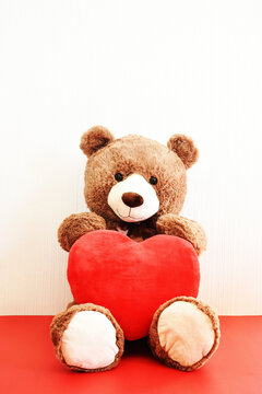 Oso de peluche caf&eacute; Con un coraz&oacute;n rojo sobre las piernas en un fondo blanco y rojo, espacio para texto en la parte superior. Dia de San Valent&iacute;n.