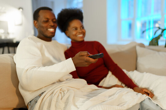 Cute Loving Couple Rest At Home On Couch. African Man Changing Channels On Tv Using Remote Control Embrace Black Woman Happy Smiling. Young Family Of Two Enjoy Spending Time Together. Selective Focus